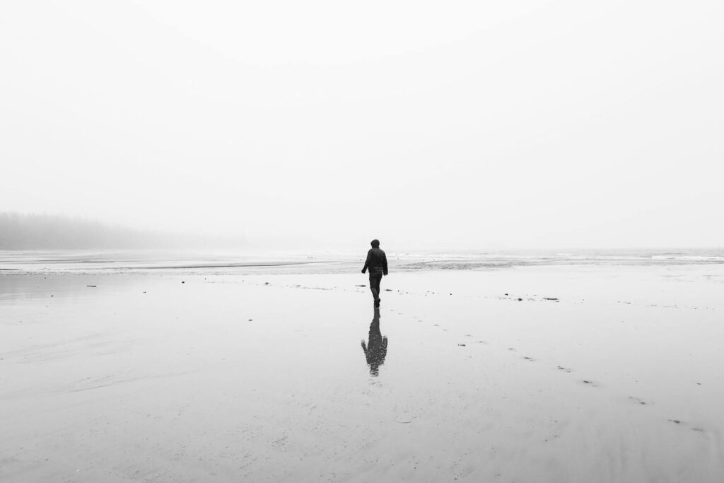 Monochrome back view of full body unrecognizable person in autumn clothes walking on lonely wet sandy coast in overcast day