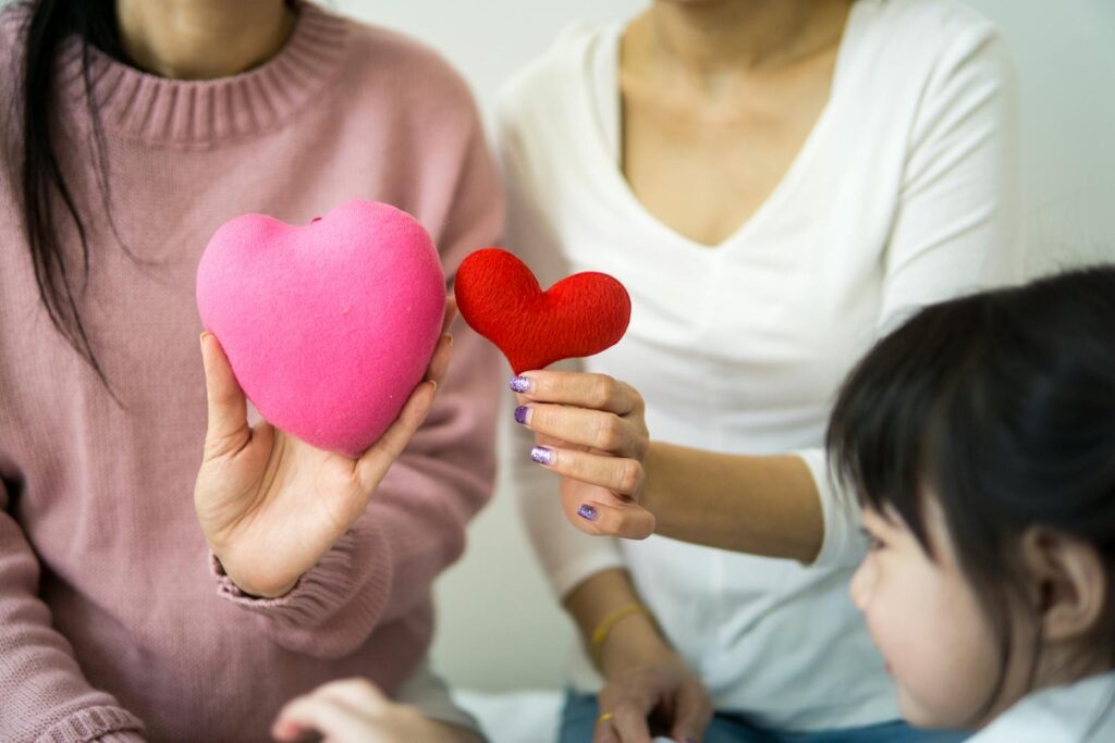 Unrecognizable ladies in casual wear demonstrating decorative hearts while sitting on couch near small kid in light room in daylight